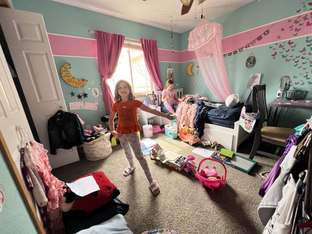 Mariana standing in her blue room with a pink stripe, filled with a bed, table, desk, and toys everywhere.