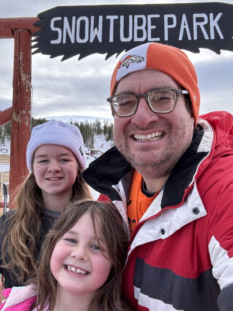 Clara, Jeromey, and Mariana, dressed in winter gear in front of a sign that says "SnowTube Park".