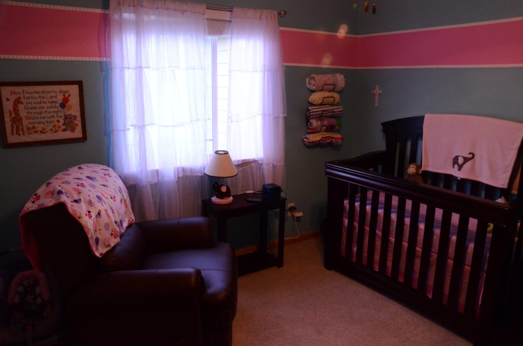 A baby nursery with blue walls, a pink stripe on the salls, a brown chair and a wooden crib.